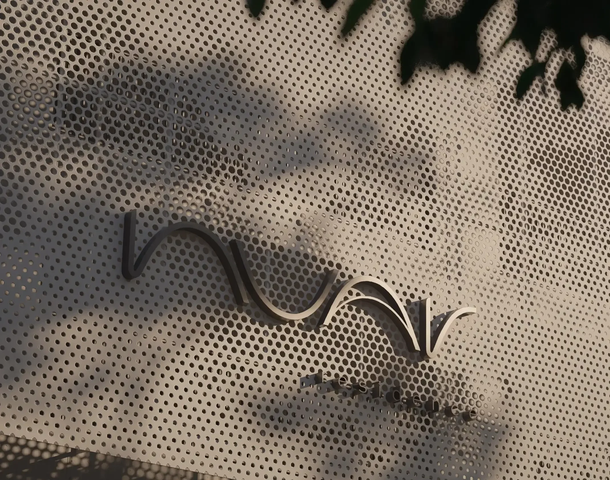 Logotipo Nuar Residence em relevo metálico sobre painel perfurado da fachada do edifício, com sombras de árvores projetadas ao fundo
